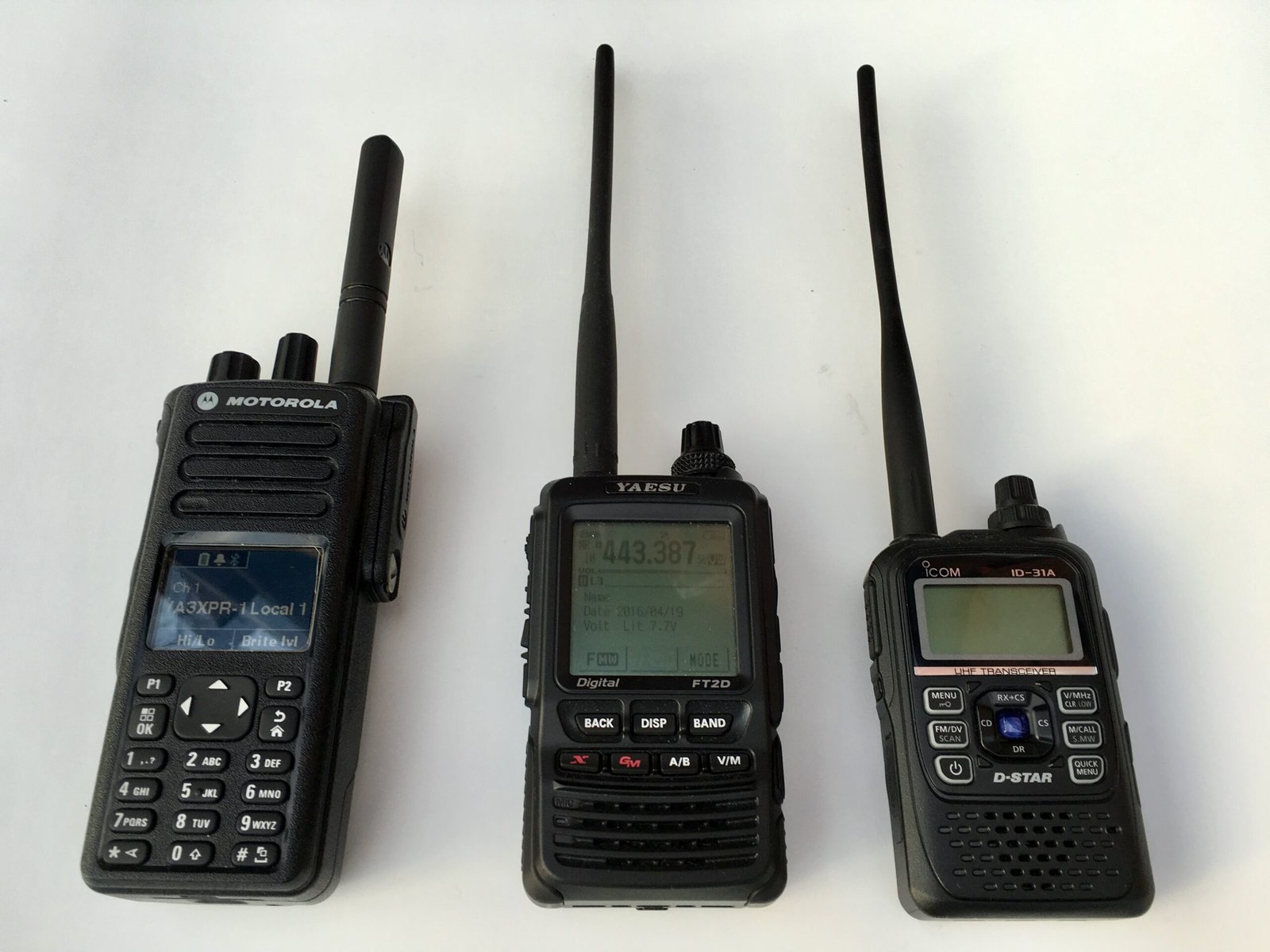 DMR vs Fusion vs D-Star handheld ham radios side-by-side: Motorola XPR7550 (DMR), Yaesu FT2D (Fusion), and Icom ID-31A (D-Star) on a white background.