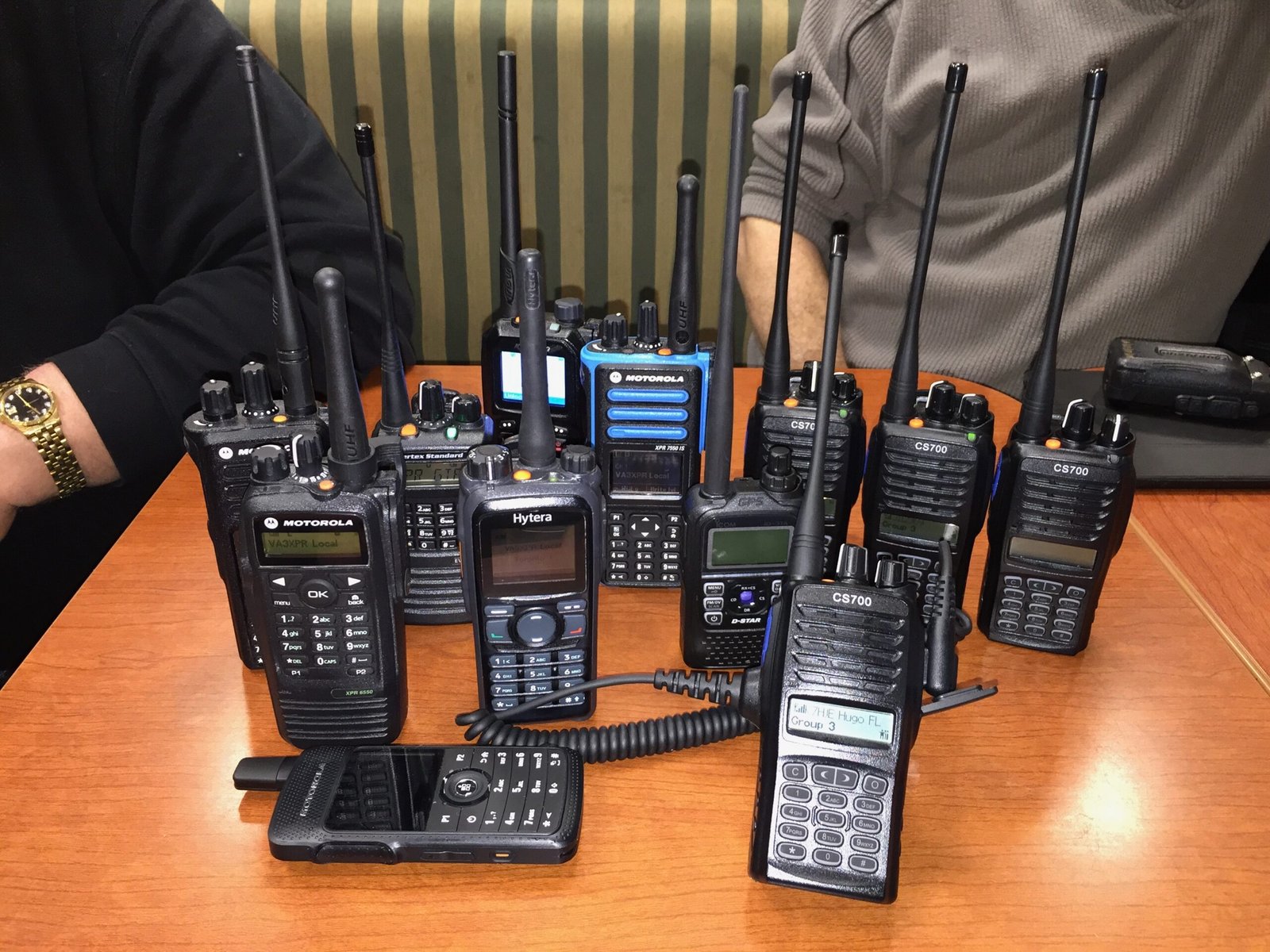 Table full of handheld digital voice ham radios at a club meeting, featuring various brands and models supporting DMR vs Fusion vs D-Star modes.